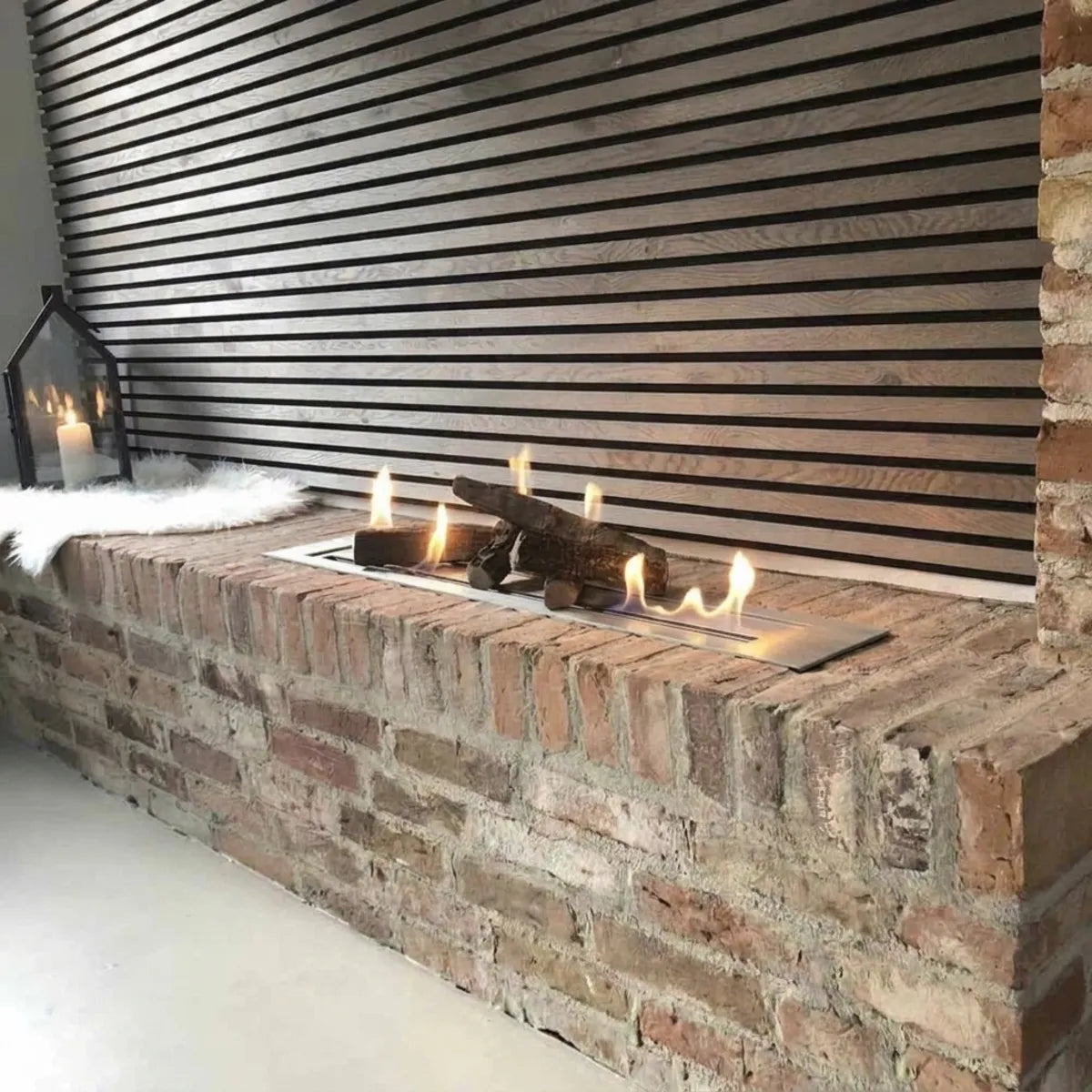 Superior 6,0L ethanol burner 120 cm with decorative logs and lit flames on a brick ledge in front of a wooden slat wall